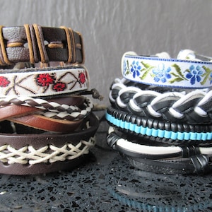 Op de afbeelding: Een set van vijf leren armbanden met gevlochten en geweven ontwerpen. Een armband heeft een witte band met rode bloemenborduurwerk, een andere heeft een witte band met blauwe bloemenborduurwerk. De armbanden zijn gestapeld op een donker oppervlak.