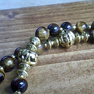 K&ouml;nnte beinhalten: Ein doppelreihiges Armband mit braunen und goldenen Perlen. Die Perlen sind aus Tigerauge und haben eine goldene Metallic-Oberfl&auml;che. Das Armband hat einen dekorativen Verschluss.