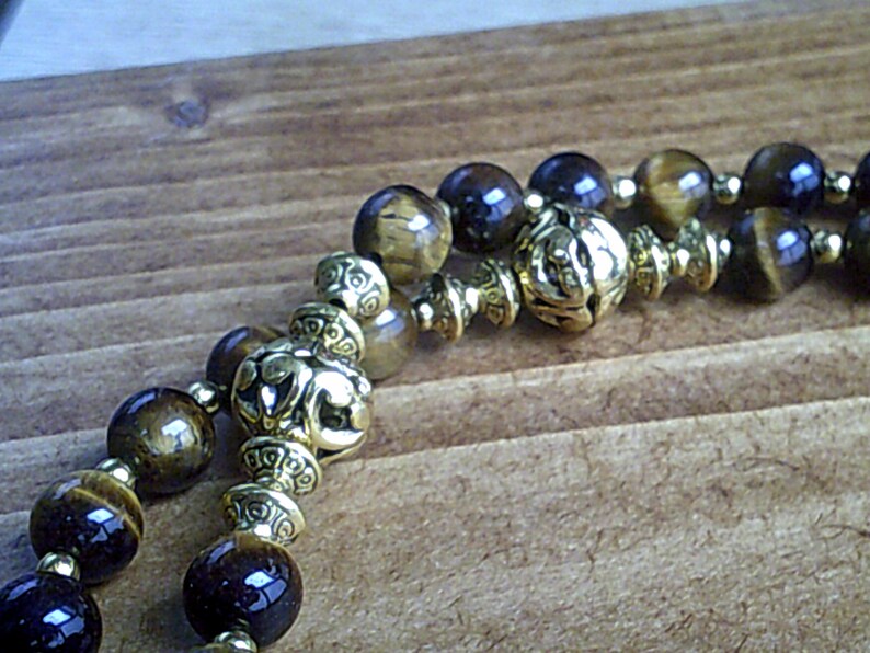 K&ouml;nnte beinhalten: Ein doppelreihiges Armband mit braunen und goldenen Perlen. Die Perlen sind aus Tigerauge und haben eine goldene Metallic-Oberfl&auml;che. Das Armband hat einen dekorativen Verschluss.