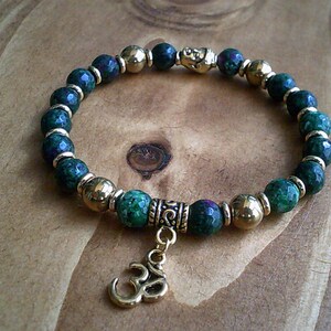 Peut inclure: Un bracelet vert et or avec un charme représentant le symbole Om. Le bracelet est composé de perles de pierres précieuses vertes et de perles de métal doré.