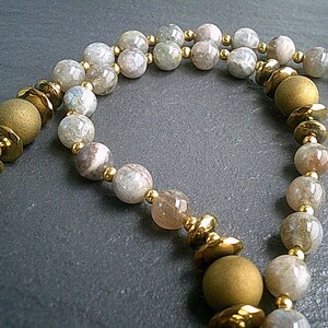 May include: A necklace made of light grey and brown gemstone beads with gold accents. The necklace has a long, flowing design and is perfect for adding a touch of elegance to any outfit.