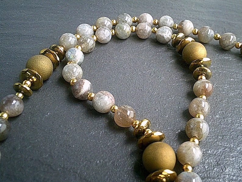 May include: A necklace made of light grey and brown gemstone beads with gold accents. The necklace has a long, flowing design and is perfect for adding a touch of elegance to any outfit.