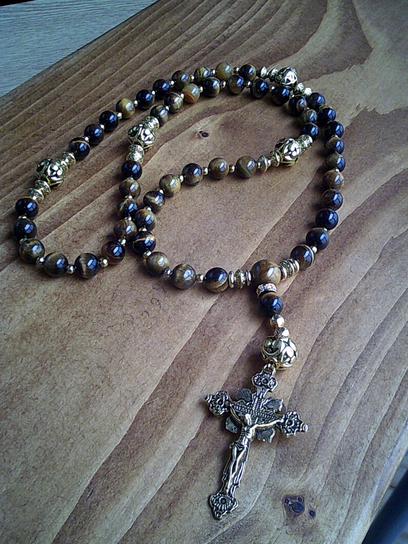 K&ouml;nnte beinhalten: Eine braune und schwarze Tigerauge-Rosenkranzperlenkette mit goldenen Akzenten und einem goldenen Kruzifix.