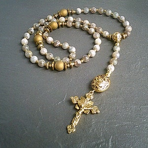 May include: A gold and white beaded rosary with a gold cross and a gold centrepiece. The beads are made of a light-coloured stone with a speckled pattern.