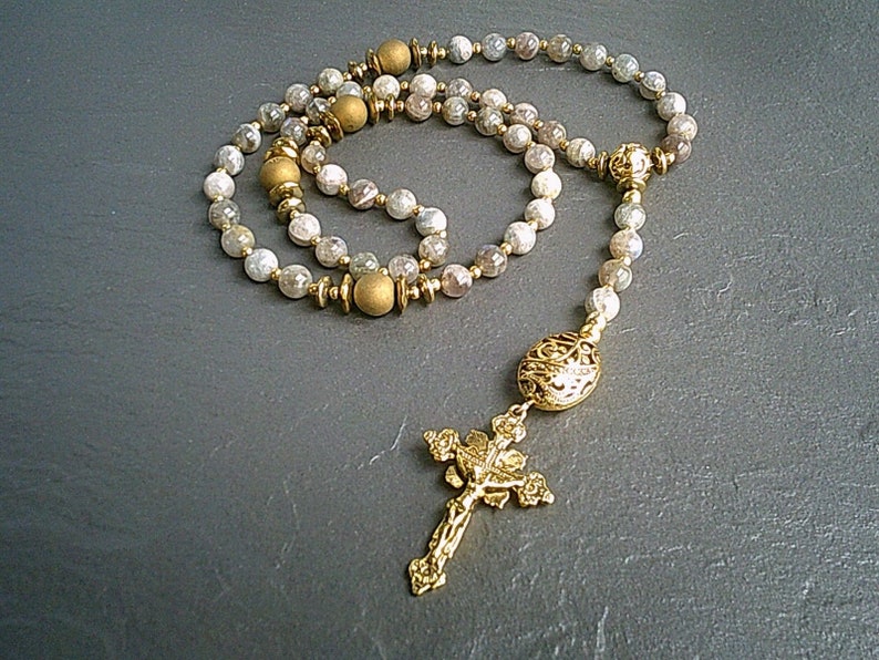 May include: A gold and white beaded rosary with a gold cross and a gold centrepiece. The beads are made of a light-coloured stone with a speckled pattern.