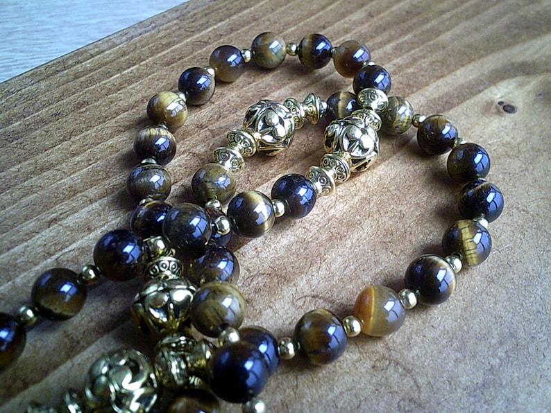 K&ouml;nnte beinhalten: Eine braune und goldene Perlenkette mit dekorativen goldenen Akzenten. Die Perlen sind aus Tigerauge-Stein.