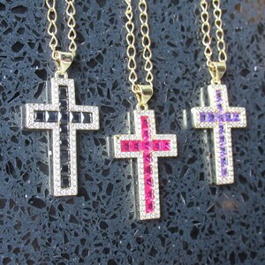 May include: Three gold-coloured cross pendants with gemstone inlays. One cross has black stones, another pink, and the third purple. Each cross is outlined with clear stones and hangs from a gold chain.