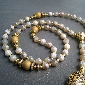 May include: A long strand of grey and white gemstone beads with gold accents and a gold pendant. The beads are strung on a gold chain.
