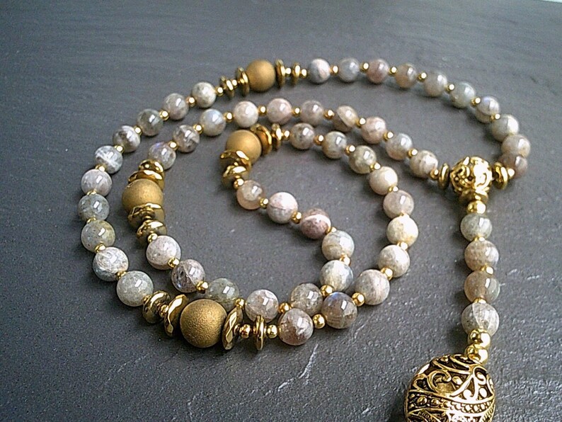 May include: A long strand of grey and white gemstone beads with gold accents and a gold pendant. The beads are strung on a gold chain.