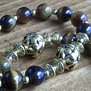 K&ouml;nnte beinhalten: Ein braunes und goldenes Perlenarmband mit dekorativen Metallakzenten. Die Perlen sind eine Mischung aus Tigerauge und dunkelblauen Steinen.