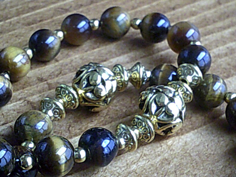 K&ouml;nnte beinhalten: Ein braunes und goldenes Perlenarmband mit dekorativen Metallakzenten. Die Perlen sind eine Mischung aus Tigerauge und dunkelblauen Steinen.