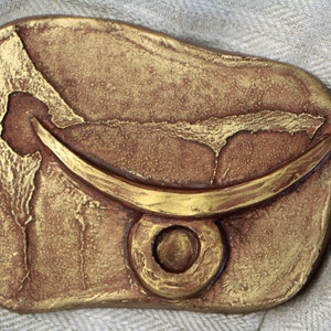 May include: A gold-coloured, textured, ceramic wall hanging with a crescent moon design and a circle in the centre.