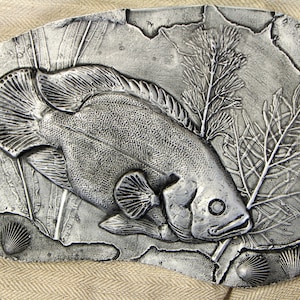 May include: A silver metal wall art piece depicting a fish with seaweed and seashells. The fish is in relief and has a textured surface.