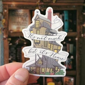 May include: A white sticker with a cartoon illustration of a crooked house with a banner that reads "It's not much, but it's home".
