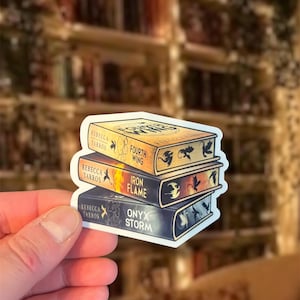 Pode incluir: Um adesivo recortado apresentando uma pilha de três livros de Rebecca Yarros. O livro superior é Fourth Wing em dourado, o do meio é Iron Flame em laranja e amarelo, e o inferior é Onyx Storm em azul escuro, todos com ilustrações de dragões.