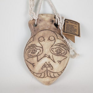 Könnte beinhalten: Ein hängender Keramik-Anhänger mit einer Schnuröse. Der Anhänger ist mit einem braunen und weißen Design eines Vogels mit geometrischen Mustern verziert. Der Anhänger hat ein Etikett mit der Aufschrift "Nuestro Barro" und Informationen über das Produkt.