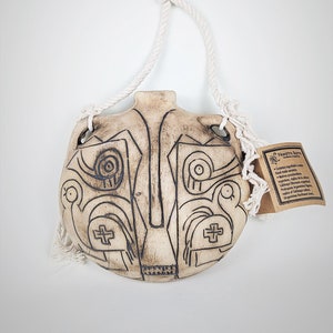 Könnte beinhalten: Ein Keramik-Wandbehang mit einem abstrakten Schwarz-Weiß-Design. Der Behang ist an einem weißen Seil befestigt und hat ein kleines braunes Etikett mit dem Text "Nuestro Barrio" und Informationen über das Produkt.