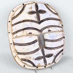 May include: Oval, off-white mask with black and blue accents. The mask features a central rectangular nose and four cross-shaped designs. Horizontal curved lines and small holes around the edge add detail. The mask is displayed on a clear stand.