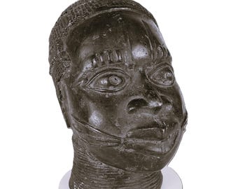 May include: A dark bronze sculpture of a human head with detailed facial features, including large eyes, a prominent nose, and a defined mouth. The head has a textured surface and a detailed hairstyle. The sculpture is mounted on a white base.