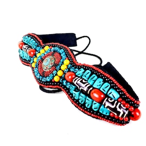 May include: A handcrafted headband featuring a vibrant array of turquoise, red, yellow, and black beads. The central design includes a circular, ornate metal piece. The headband has a black suede backing and black ties for securing.
