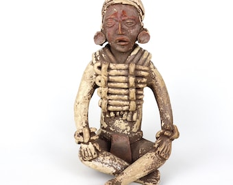 May include: A ceramic figurine of a seated figure with a headdress, wearing a decorative chest piece and large earrings. The figurine is painted in shades of brown, red, and white.