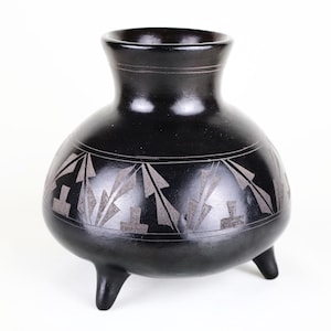 May include: A black ceramic vase with a rounded body and three short legs. The vase features a narrow neck and a decorative band of geometric patterns in a lighter color around the midsection. The vase is likely handmade.