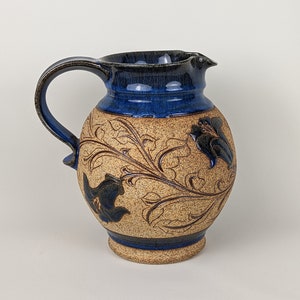 May include: A brown ceramic pitcher with a blue glaze and floral design. The pitcher has a wide base and a handle.