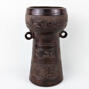 Può includere: Un vaso decorativo marrone scuro con una parte superiore ampia e una base svasata. Il vaso presenta intricati disegni in rilievo, tra cui figure e fogliame. Due manici circolari sono sulla parte superiore.