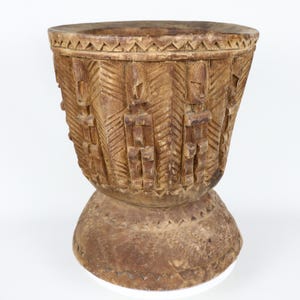 May include: A carved wooden vessel with a wide top and a pedestal base. The exterior features intricate geometric and figurative carvings, with a natural wood finish. The vessel's design suggests a cultural or tribal origin.