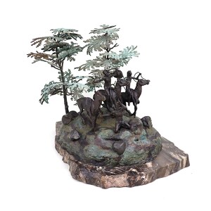 May include: A bronze sculpture depicting a group of Native American figures on horseback, hunting a deer. The sculpture is set on a base of green and brown stone, with two trees made of metal leaves.