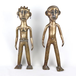 May include: Two antique bronze figurines of human figures. The figures have elongated features, with detailed faces and simple, stylized bodies. One figure has a crown-like headpiece, and the other has a simple cap. Both figures stand with their arms at their sides.