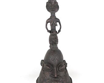 May include: A bronze-colored bell-shaped sculpture with a detailed face at the base and a series of figures stacked above. The top figure has a tall, pointed head. The sculpture is set against a white background.
