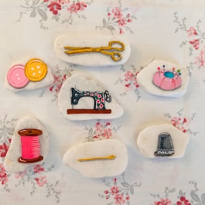 May include: Seven white painted rocks with various sewing-themed designs. The rocks feature a sewing machine, a spool of thread, a thimble, a needle, a pair of scissors, and two buttons.