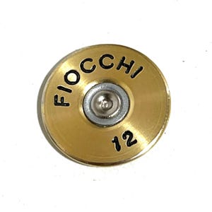 May include: A brass-colored, circular object with the word "FIOCCHI" in black lettering and the number "12". The center features a silver-colored, circular component. The object is likely a component or decorative element.