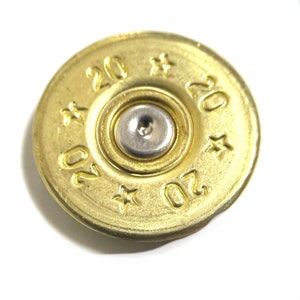 May include: Close-up of a brass-colored object with the number "20" stamped four times around the edge. Small star shapes are also stamped around the edge. A silver-colored metal piece is in the center.