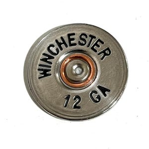 May include: A silver-colored Winchester 12 GA shotgun shell casing. The word "WINCHESTER" is arched across the top, and "12 GA" is printed at the bottom. The center has a copper-colored ring.