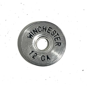 May include: A silver-colored Winchester 12 GA shotgun shell casing. The text "WINCHESTER" is arched across the top, and "12 GA" is printed at the bottom. The casing has a circular shape with a central hole.