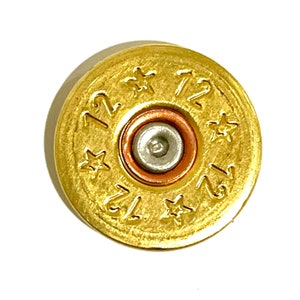 May include: Close-up of a gold-colored 12-gauge shotgun shell casing. The casing has the number "12" repeated three times, along with star-shaped designs. The center features a silver primer surrounded by a copper ring.