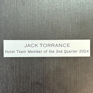 May include: A white rectangular sign with black text that reads "JACK TORRANCE Hotel Team Member of the 2nd Quarter 2024".