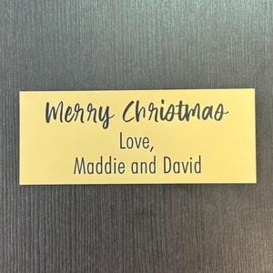 May include: A gold-colored rectangular sign with the text "Merry Christmas - Love, Maddie and David" written in black.