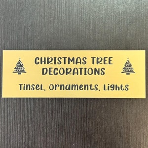 May include: A gold-colored metal sign with black text and images of Christmas trees. The sign reads "Christmas Tree Decorations" and "Tinsel, ornaments, lights".