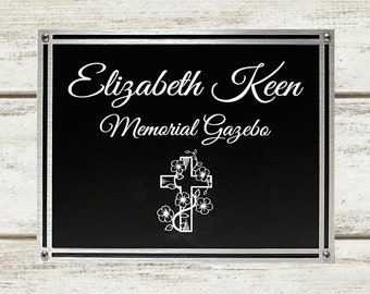 May include: Black metal sign with silver trim and rounded corners. The sign reads "Elizabeth Keen Memorial Gazebo" with a stylized cross and floral design.