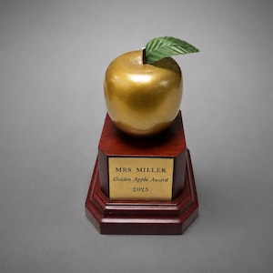 May include: A gold-colored apple trophy with a single green leaf on top, mounted on a dark brown wooden base. A gold-colored metal plaque on the front of the base features the text: MRS. MILLER, Golden Apple Award, 2025, in black lettering.