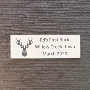 May include: A white plaque with a silhouette of a deer head with antlers. The text on the plaque reads "Ed's First Buck, Willow Creek, Iowa, March 2020".