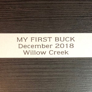 May include: An off-white rectangular label with the text "MY FIRST BUCK December 2018 Willow Creek" in brown, serif font. The label is on a dark brown, wood-grain background.