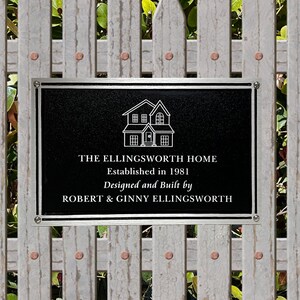 May include: A black plaque with silver trim on a white wooden fence. The plaque has a white outline of a house and the text "THE ELLINGSWORTH HOME Established in 1981 Designed and Built by ROBERT & GINNY ELLINGSWORTH"