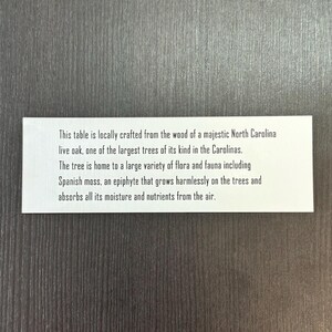 May include: A white card with black text describing a table made from the wood of a majestic North Carolina live oak. The text explains that the tree is home to a variety of flora and fauna, including Spanish moss, an epiphyte that grows harmlessly on the trees and absorbs moisture and nutrients from the air.