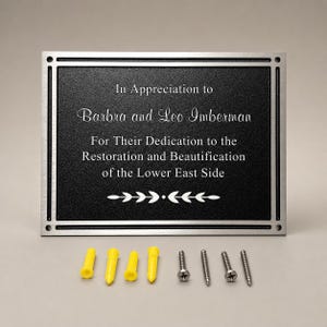 May include: A rectangular black plaque with a silver metal border, featuring the engraved text In Appreciation to Barbra and Leo Imberman For Their Dedication to the Restoration and Beautification of the Lower East Side, with four yellow anchors and four screws.