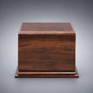 May include: A rectangular wooden box with a dark brown finish and visible wood grain. The box features a stepped base and a flat top surface with a single small, centered gold-colored circular inlay. The construction is solid and smooth.