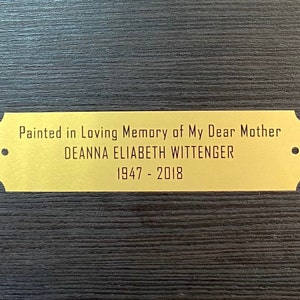 May include: A gold-colored plaque with rounded corners and two small holes. The plaque reads "Painted in Loving Memory of My Dear Mother DEANNA ELIABETH WITTENGER 1947 - 2018".